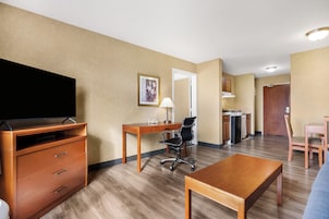 Quarto de hotel com escrivaninha de madeira, uma cadeira, televisão, cama e cozinha compacta.