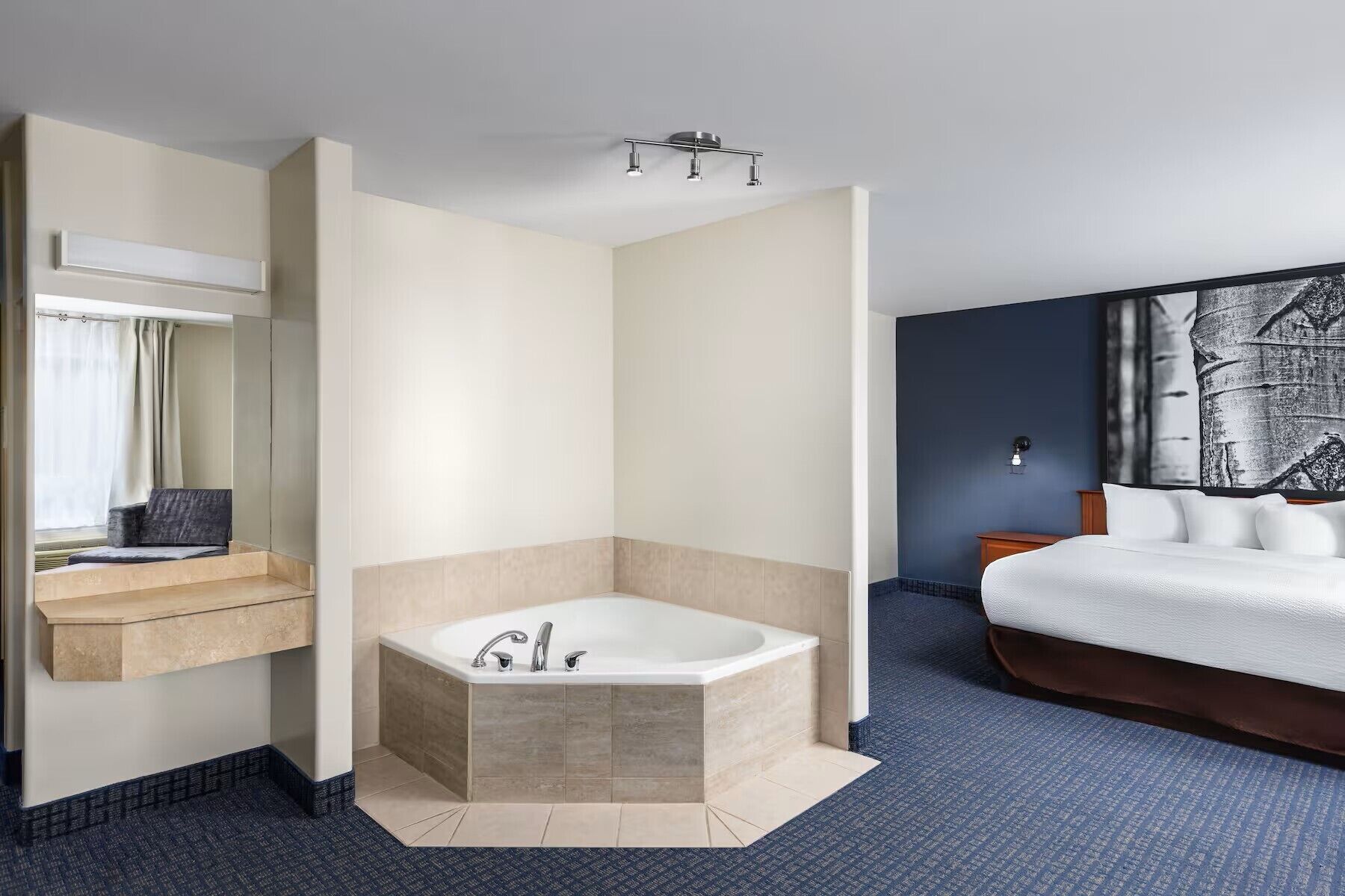 A modern hotel room with a corner bathtub, a large bed, a nightstand, a wall-mounted light, and a partial view of a bathroom with a mirror and curtains.
