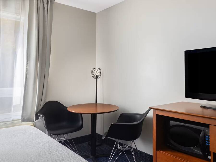 Una habitación de hotel con una cama, un televisor, un escritorio con microondas, dos sillas, una mesita y una lámpara de pie.