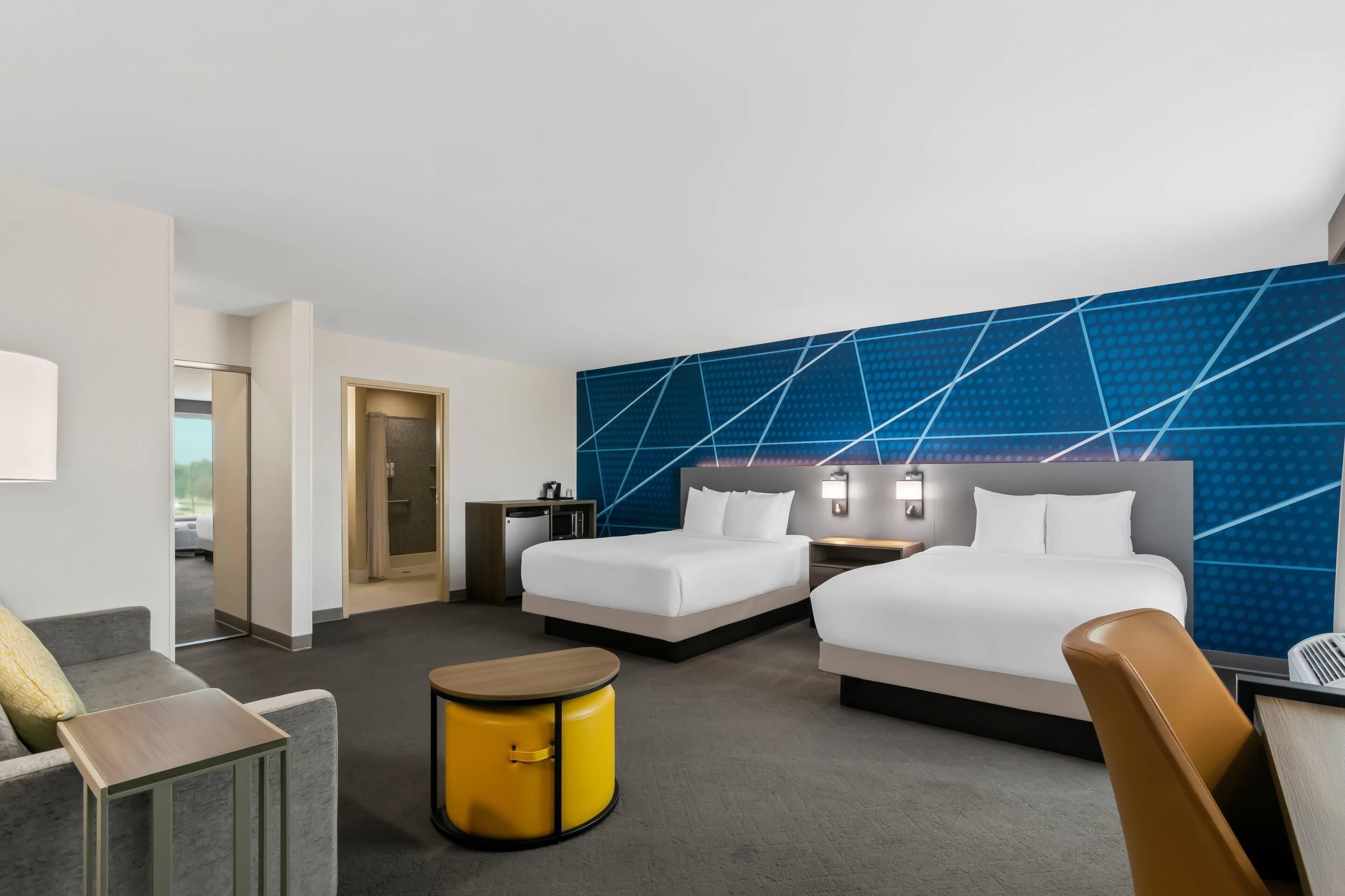 A hotel room with two beds, a desk, a chair, a small table, and a wall with a geometric pattern.