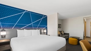 A hotel room with a large bed, a nightstand, a wall-mounted lamp, a sofa, a small table, and a yellow ottoman.