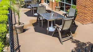 An outdoor patio with tables and chairs, a blue umbrella, and a brick building in the background.