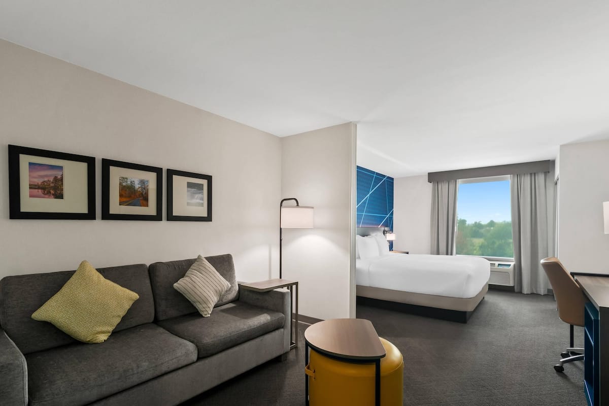 Habitación de hotel con sofá, cama, escritorio y una silla. En la pared, hay cuadros enmarcados y una ventana con cortinas.
