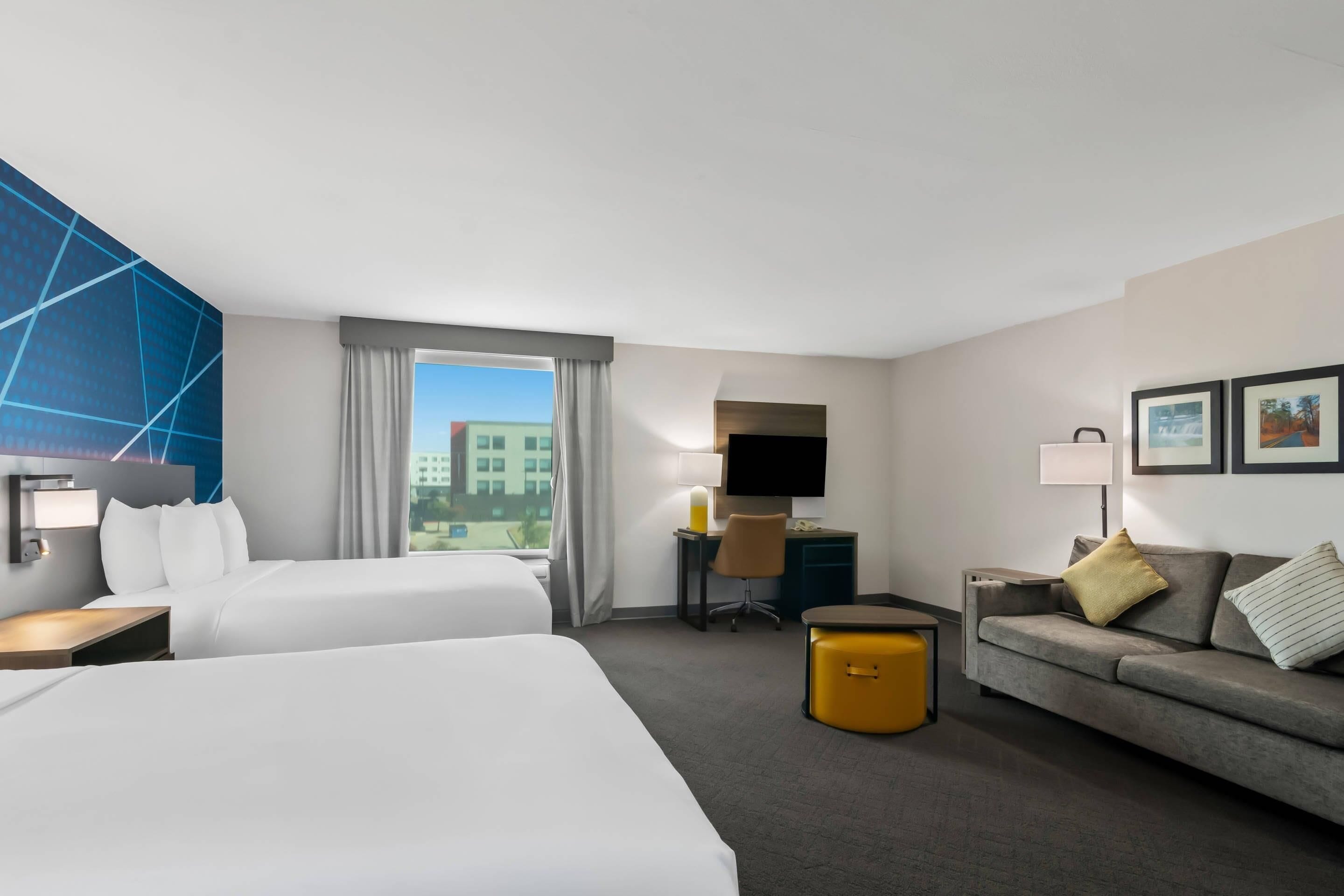 A hotel room with two beds, a sofa, a desk with a chair, a TV, and a window with curtains.