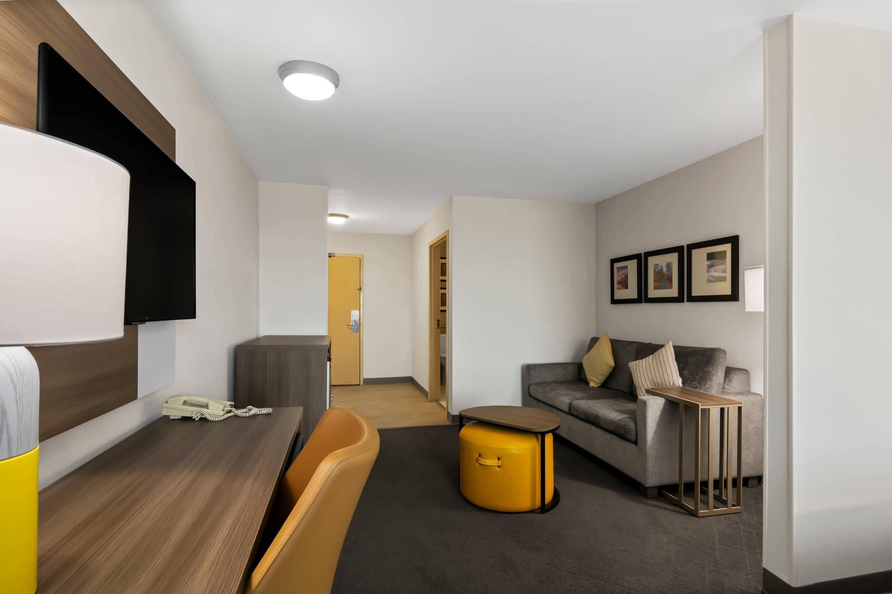 Una habitación de hotel moderna con un escritorio de madera, un sofá, una mesita pequeña y un otoman amarillo.