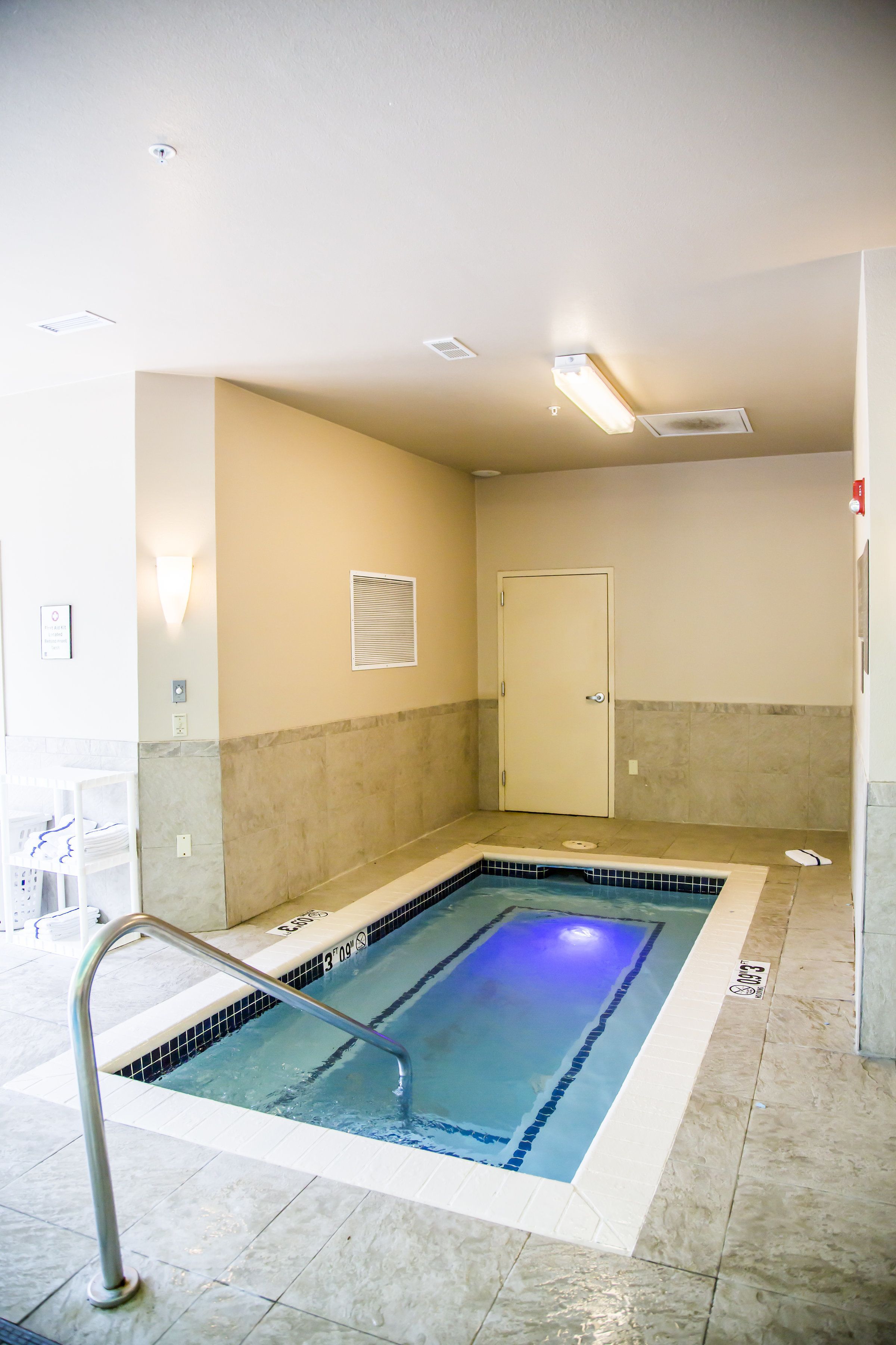 Una piscina interior pequeña con una escalera metálica, una puerta y suelo de baldosas.