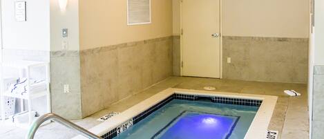 Una piscina interior pequeña con una escalera metálica, una puerta y suelo de baldosas.