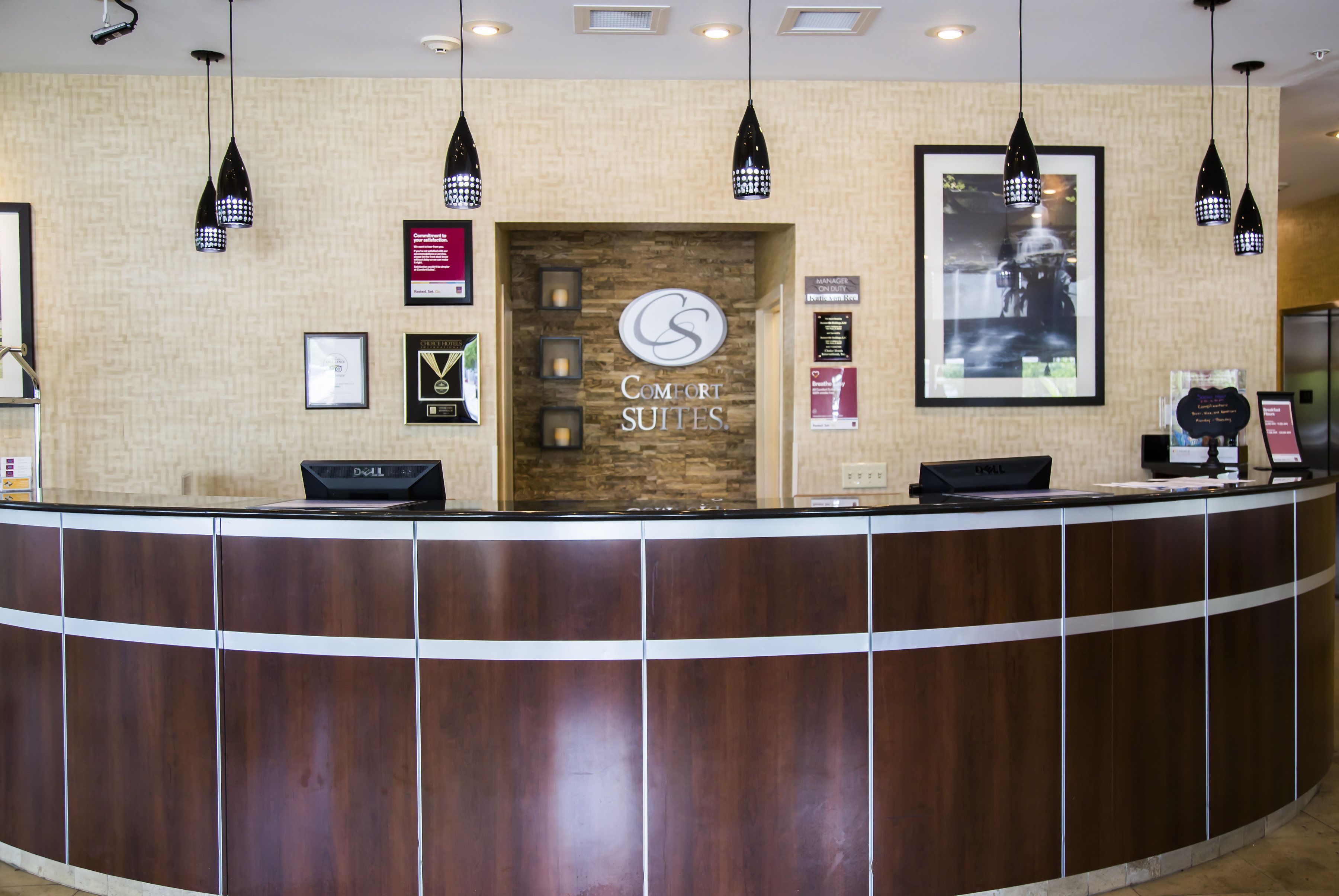 Un mostrador de recepción con detalles de madera, un cartel que dice "Comfort Suites" y varios certificados o reconocimientos enmarcados en la pared.