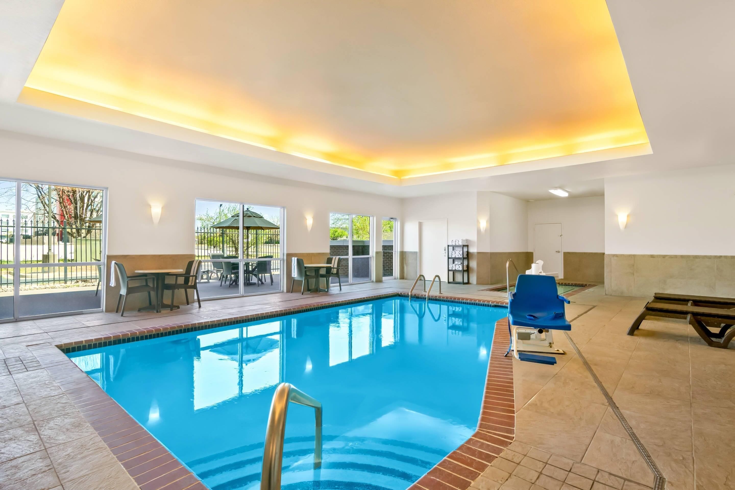 An indoor swimming pool with clear blue water, surrounded by a tiled floor and a brick border. There are lounge chairs and a table with chairs outside the pool area.