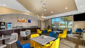 A modern cafe with yellow and blue chairs, granite tables, and a kitchen area with a coffee machine.