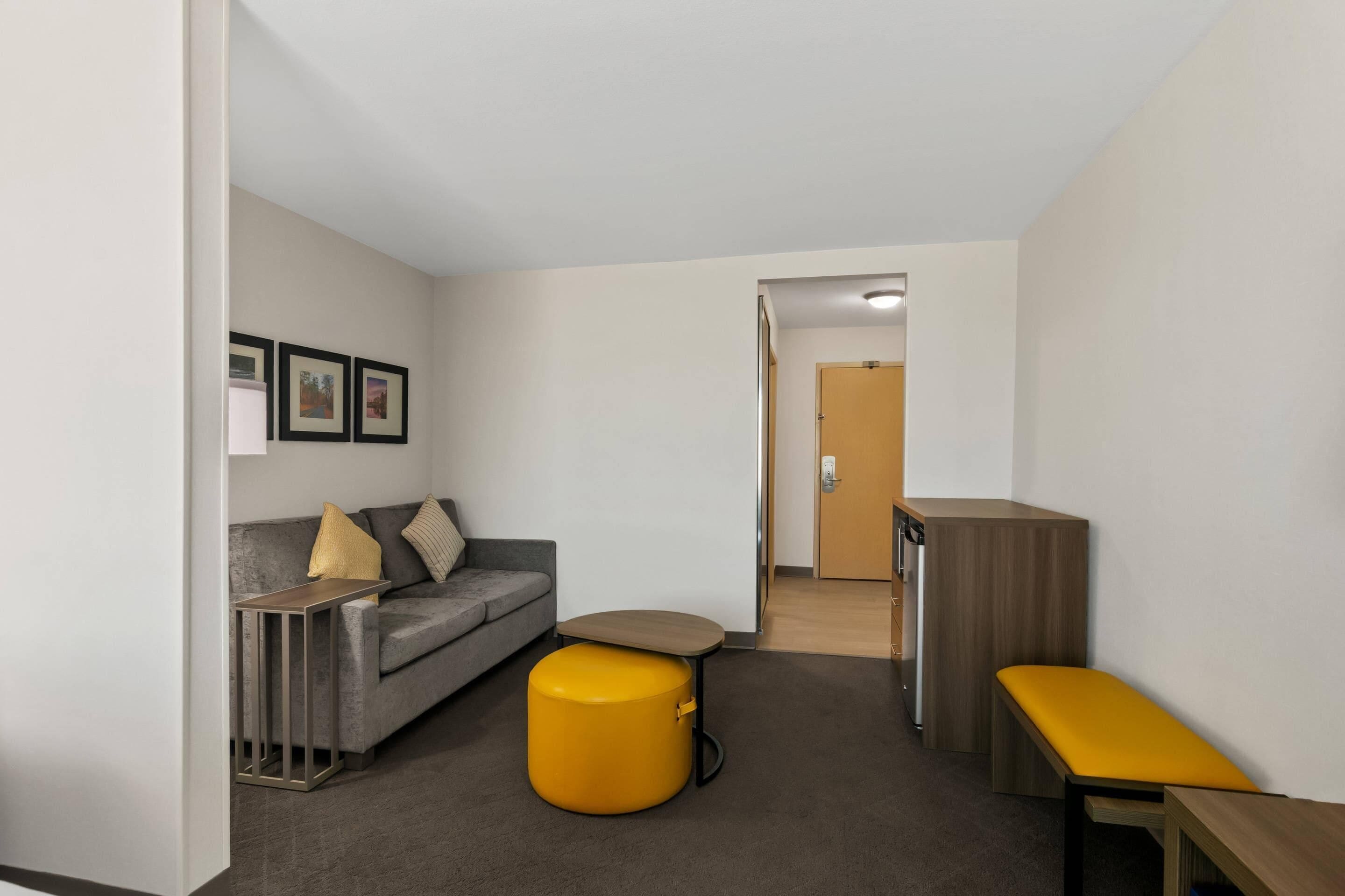 Un espace de vie moderne et compact, comprenant un canapé gris, un pouf jaune, une petite table en bois et un meuble de rangement.