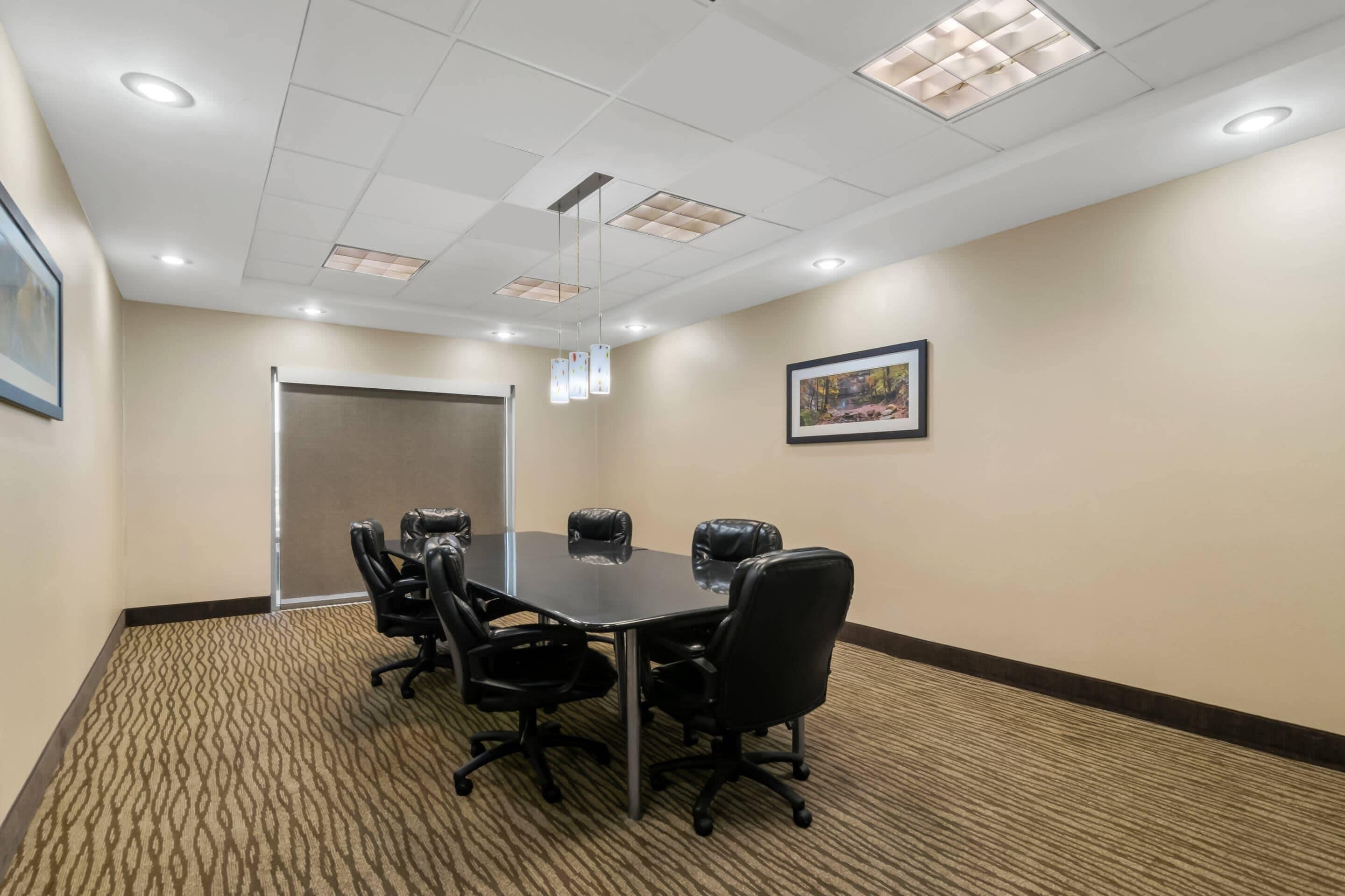 A conference room with a rectangular table, black leather chairs, recessed ceiling lights, patterned carpet, and framed artwork on the walls.