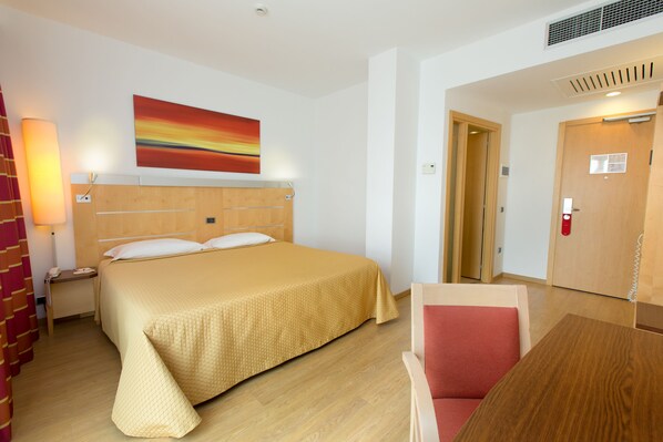 Una habitación de hotel con cama, escritorio, silla, un cuadro en la pared y una puerta.