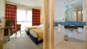 Ein Hotelzimmer mit zwei Betten, einem Schreibtisch, einem Fernseher und einem Badezimmer mit Waschbecken und Spiegel.