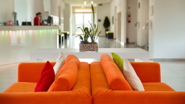 Eine helle, moderne Lobby mit einer Rezeption, einem weißen Empfangstresen und einer Sitzecke mit orangefarbenen Sofas und Kissen.