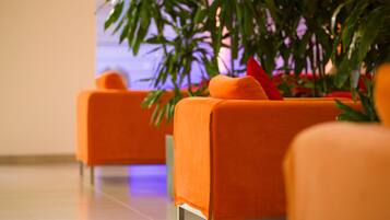 Eine Reihe von orangefarbenen Stühlen mit roten Kissen in einer modernen Lobby mit Fliesenboden und einer großen, grünen Pflanze im Hintergrund.