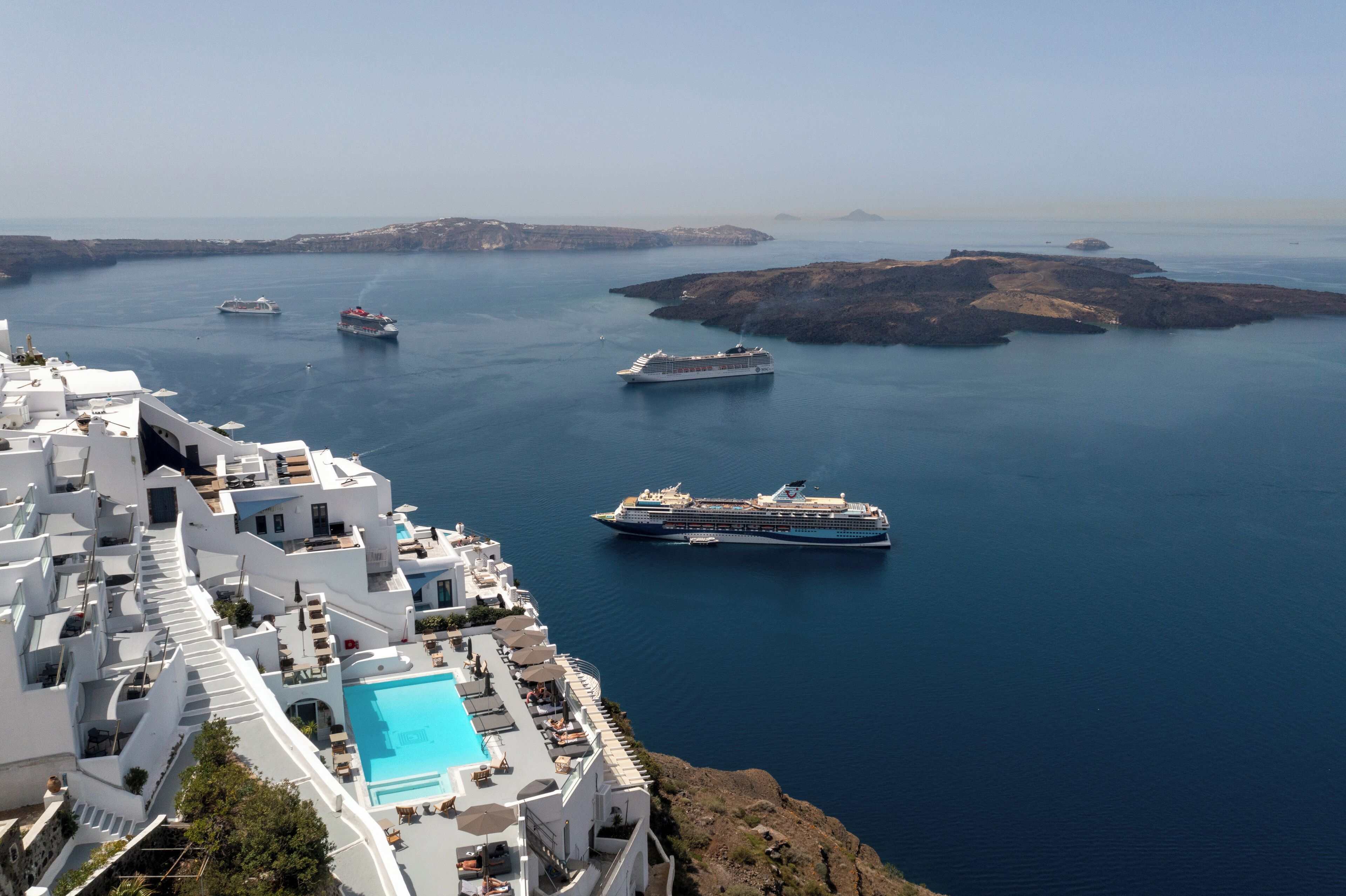 Un hotel a la costa amb piscina, diversos vaixells de creuer al fons i terreny rocos.