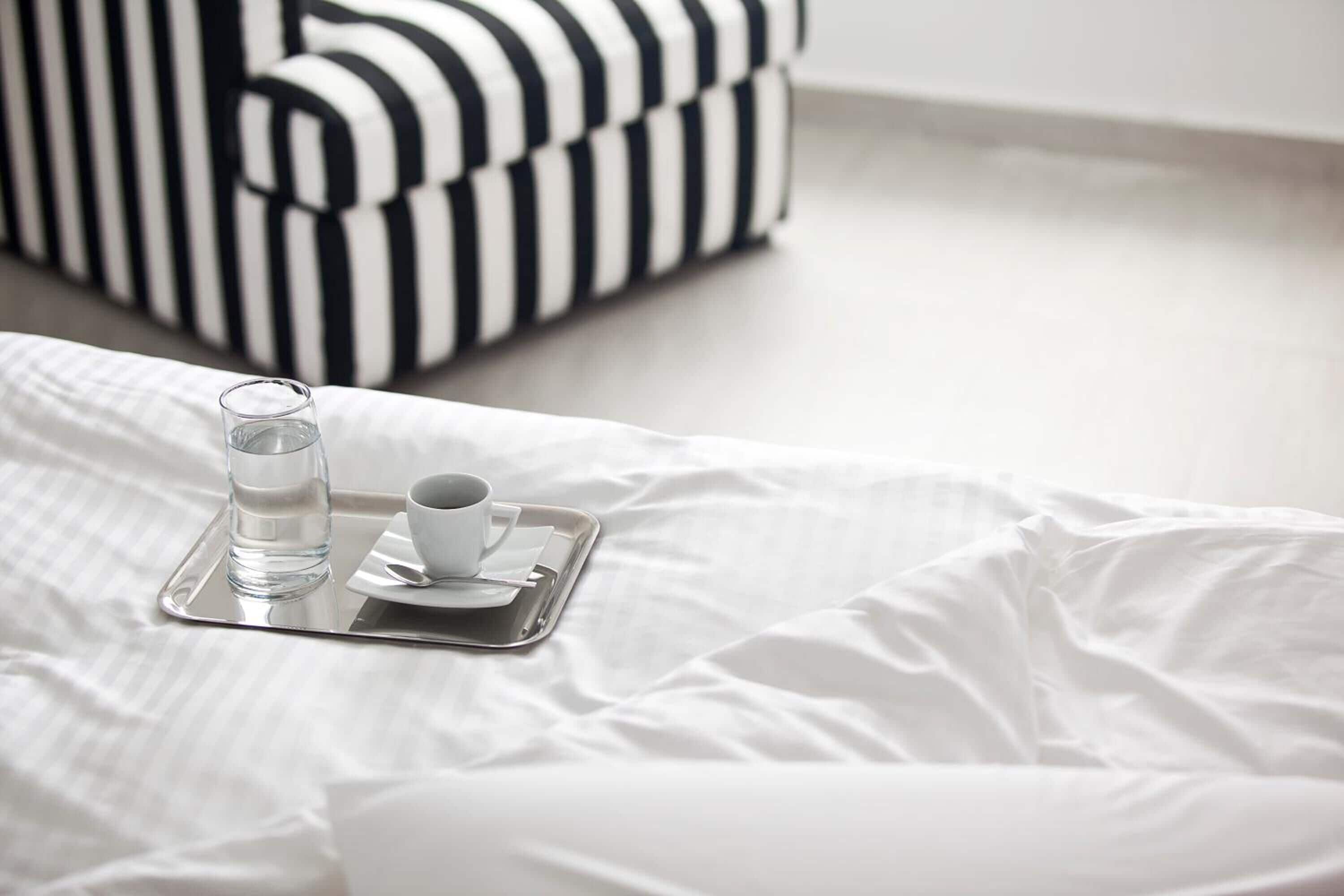 A bed with a tray containing a glass of water and a cup, a striped black and white sofa in the background, and a white bedspread.