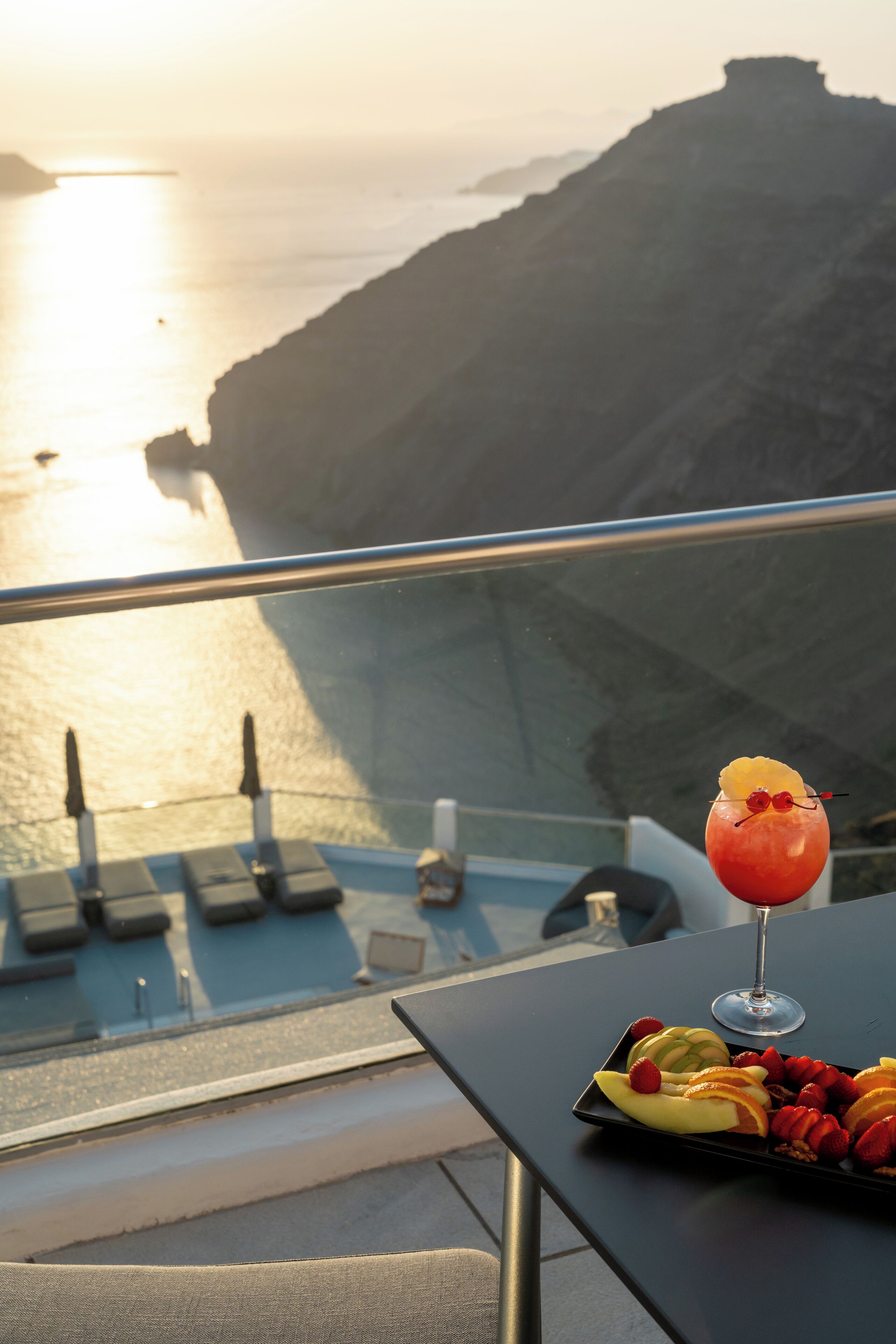 Mesa com espeto de frutas e um cocktail numa varanda com vista para a costa.