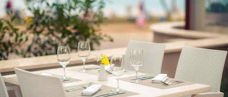 Una mesa lista para comer, con sillas blancas, un florero con flores amarillas y copas de vino.