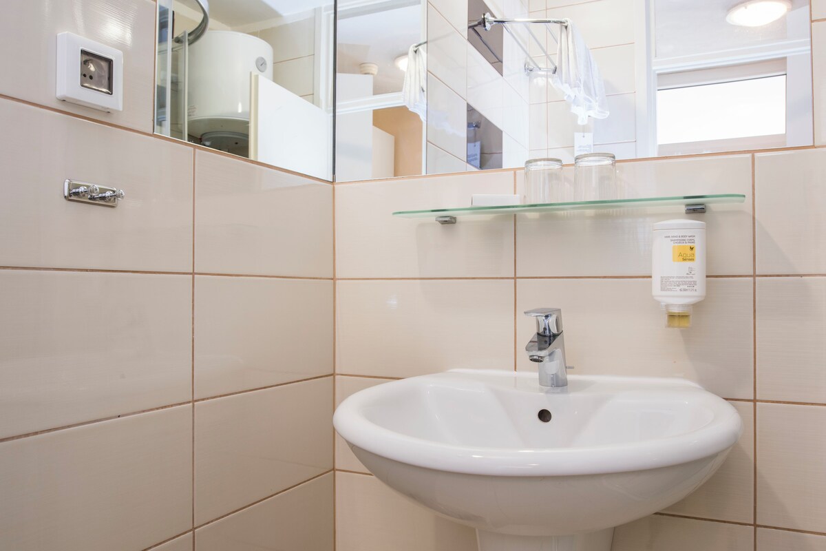 Un baño con un lavabo blanco, un estante de vidrio encima y un espejo que refleja una escalera y un reloj.