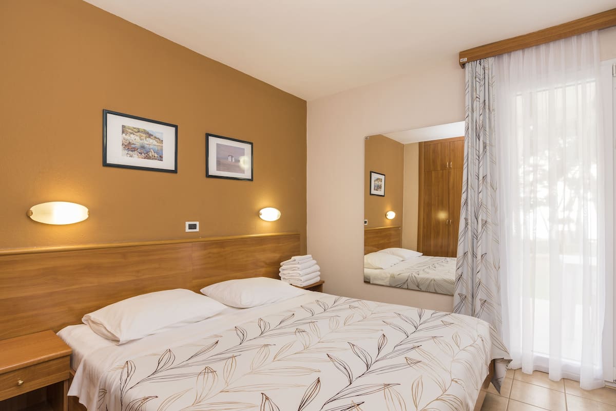 Hotellihuone, jossa on sänky, yöpöydät, seinään kiinnitetyt valaisimet, kehystetyt taulut, peili ja läpikuultavat verhot.
