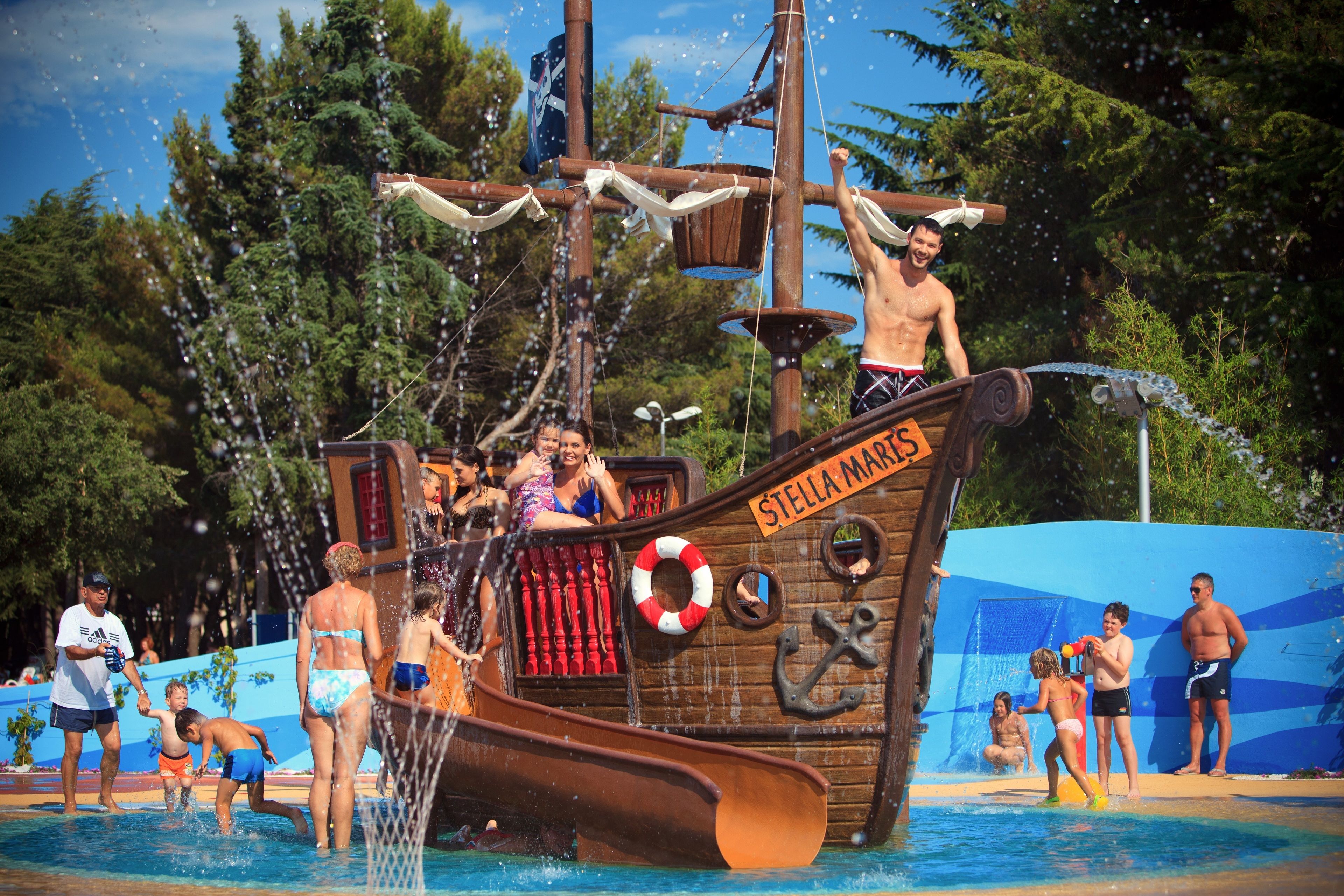A pirate ship water slide with a lifebuoy and anchor decoration, surrounded by a pool and people.