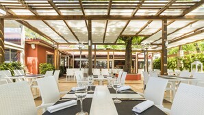 Un área de comedor al aire libre con sillas y mesas de mimbre blanco, cubierta por una pérgola con techo de vidrio.