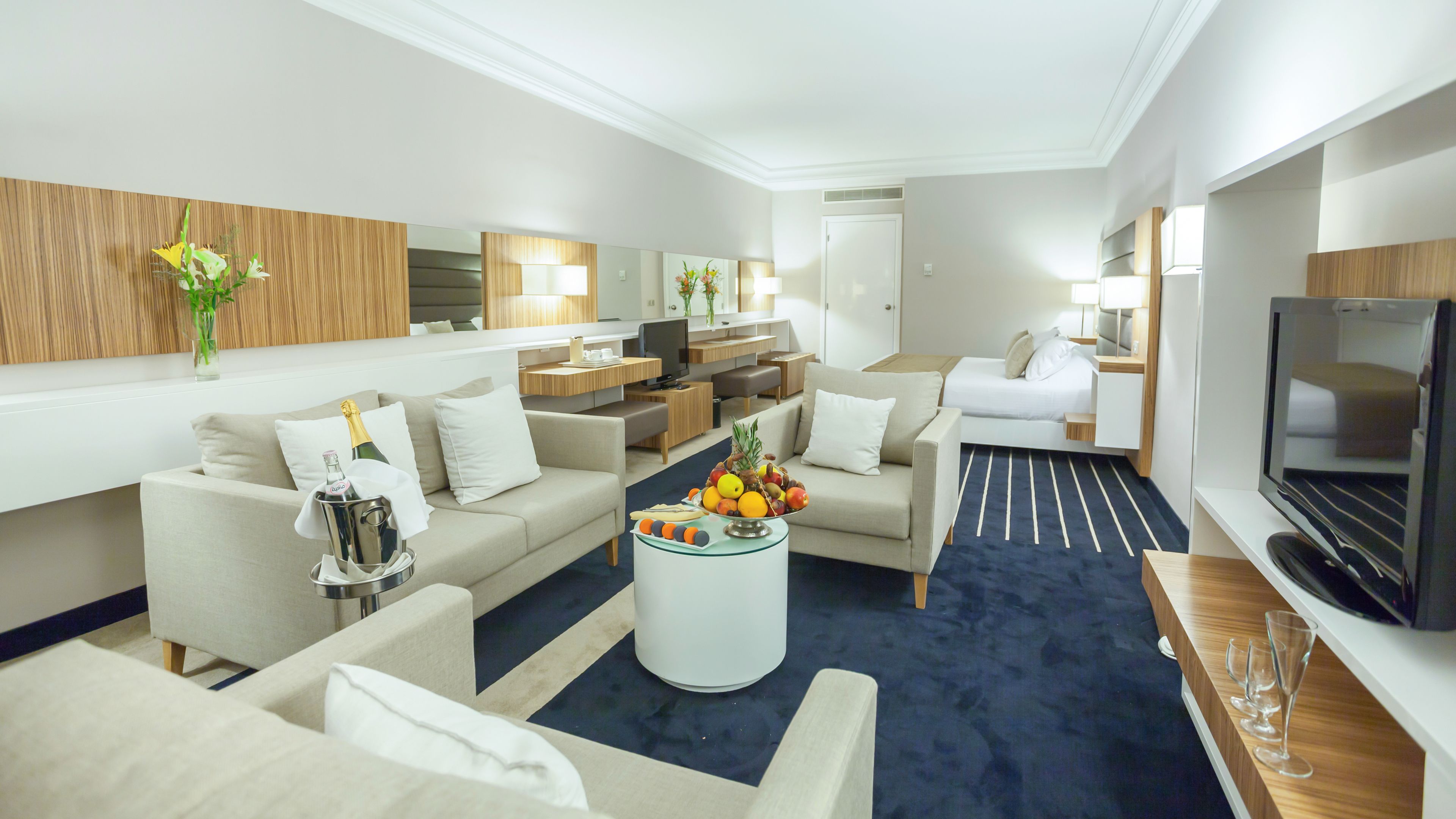 A modern hotel room with a living area, a dining area, a TV, and a minibar.