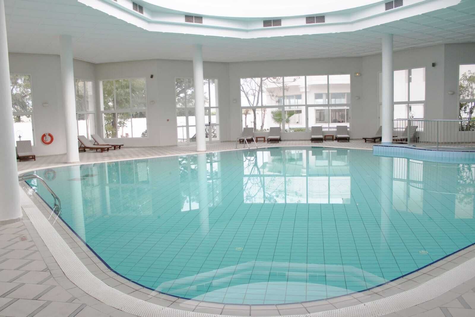 En indendørs swimmingpool med klart, blåt vand, omgivet af hvide vægge og store vinduer.