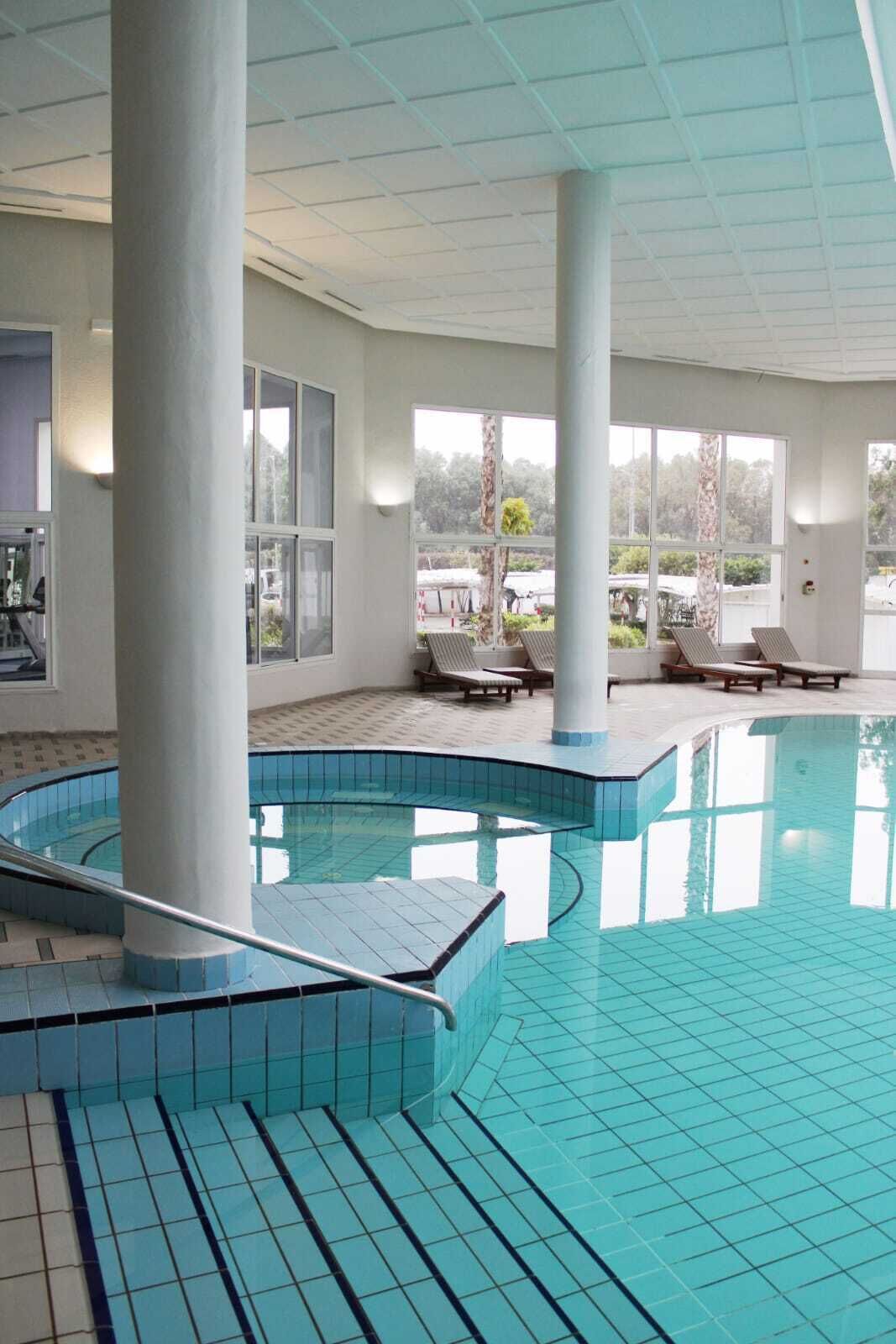 An indoor swimming pool with clear glass walls, multiple lounge chairs, and a tiled floor.