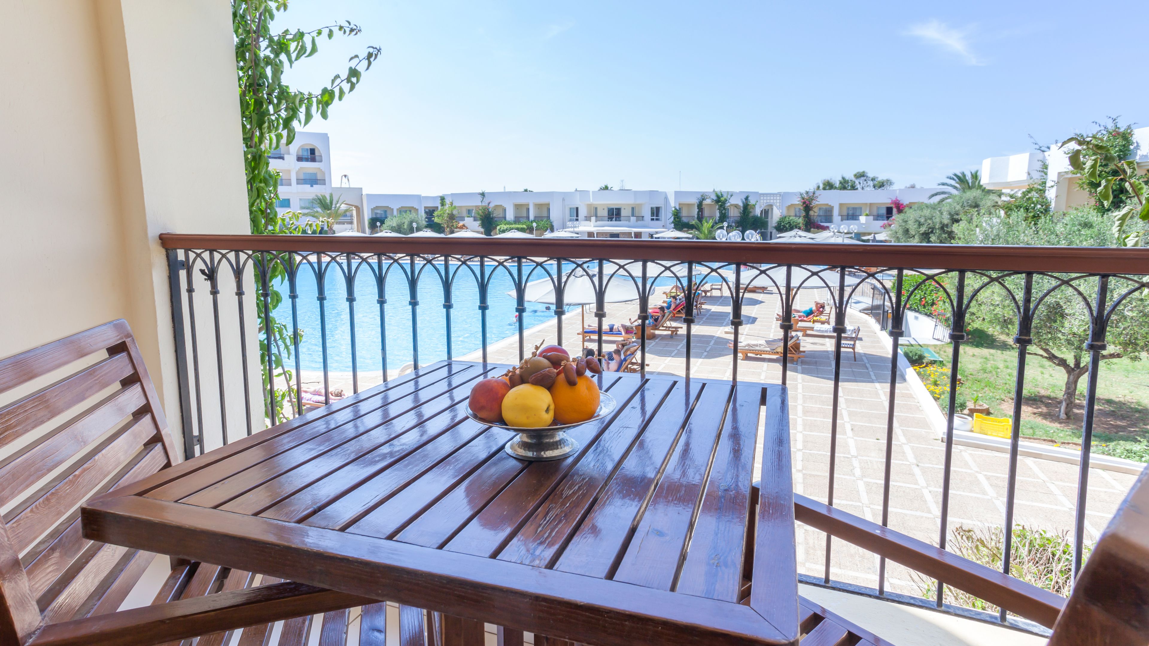 Balcón con una mesa de madera y un cuenco de fruta. Se aprecia una zona de piscina con sillones reclinables y sombrillas.