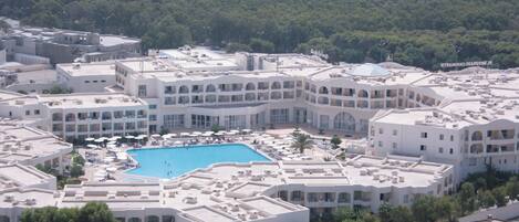 A hotel with a central swimming pool, multiple balconies, and a lush green backdrop.