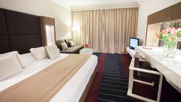 Quarto de hotel com uma cama grande, uma escrivaninha com computador, uma cadeira, uma mesa pequena com um vaso de flores e uma janela com cortinas.