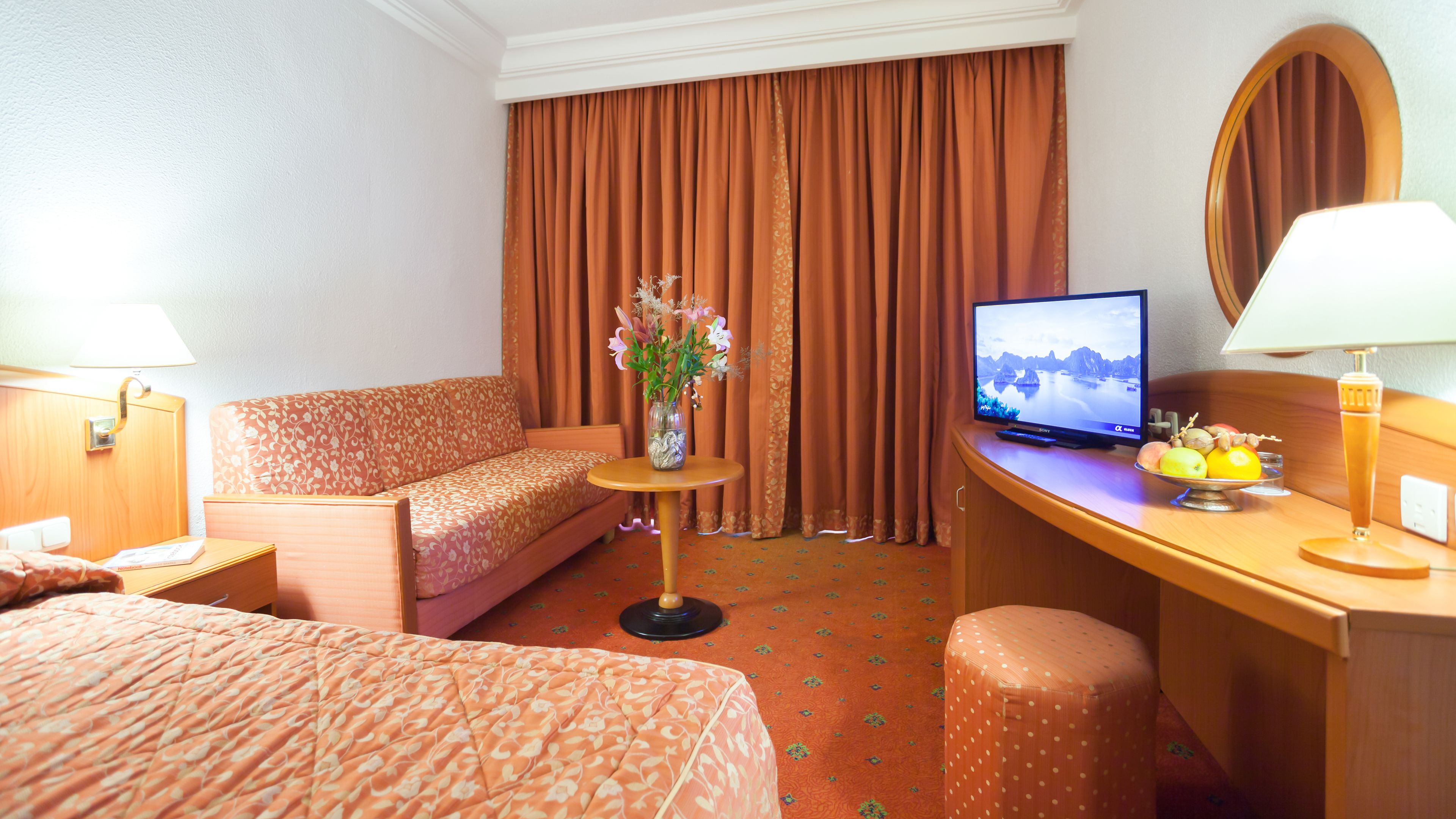 A hotel room with a bed, a sofa, a desk with a TV, a vase with flowers, and a fruit bowl.