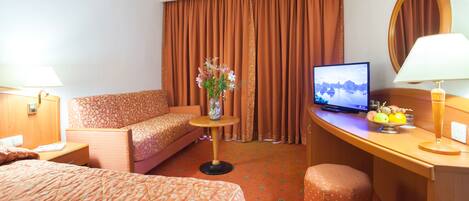A hotel room with a bed, a sofa, a desk with a TV, a vase with flowers, and a fruit bowl.