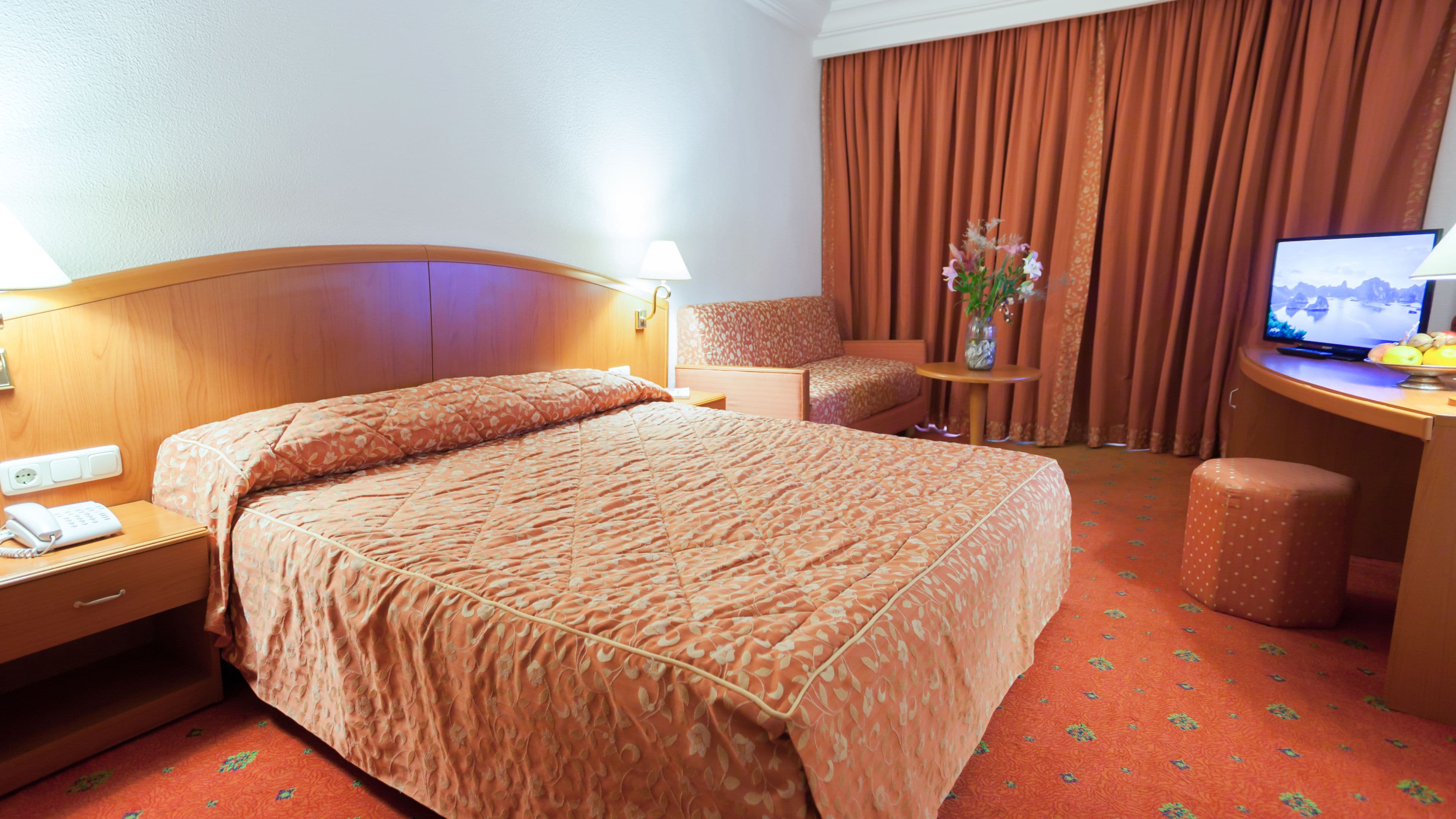 A hotel room with a large bed, a desk, a chair, a television, and a small table with a vase of flowers.