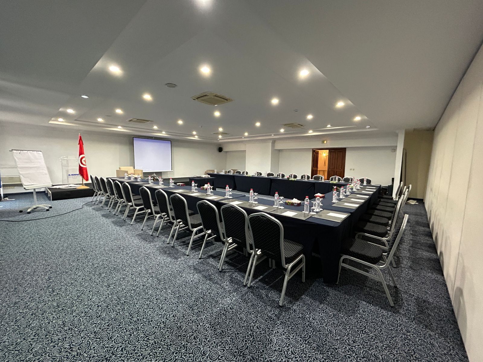 Une salle de conférence avec une longue table, des chaises, un écran de projection et un drapeau rouge.