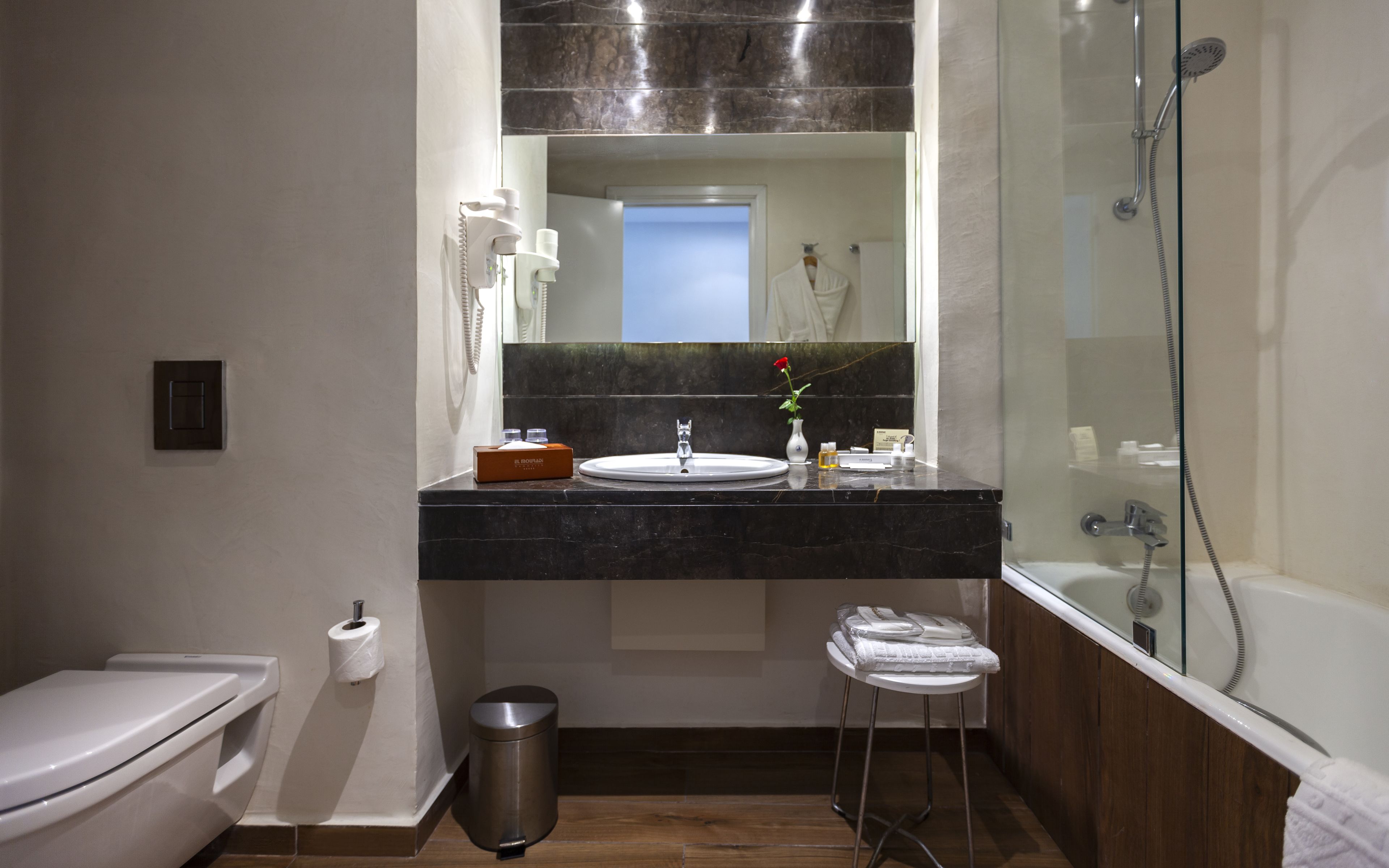 A modern bathroom with a marble countertop, a sink, a mirror, a toilet, a trash can, and a shower.