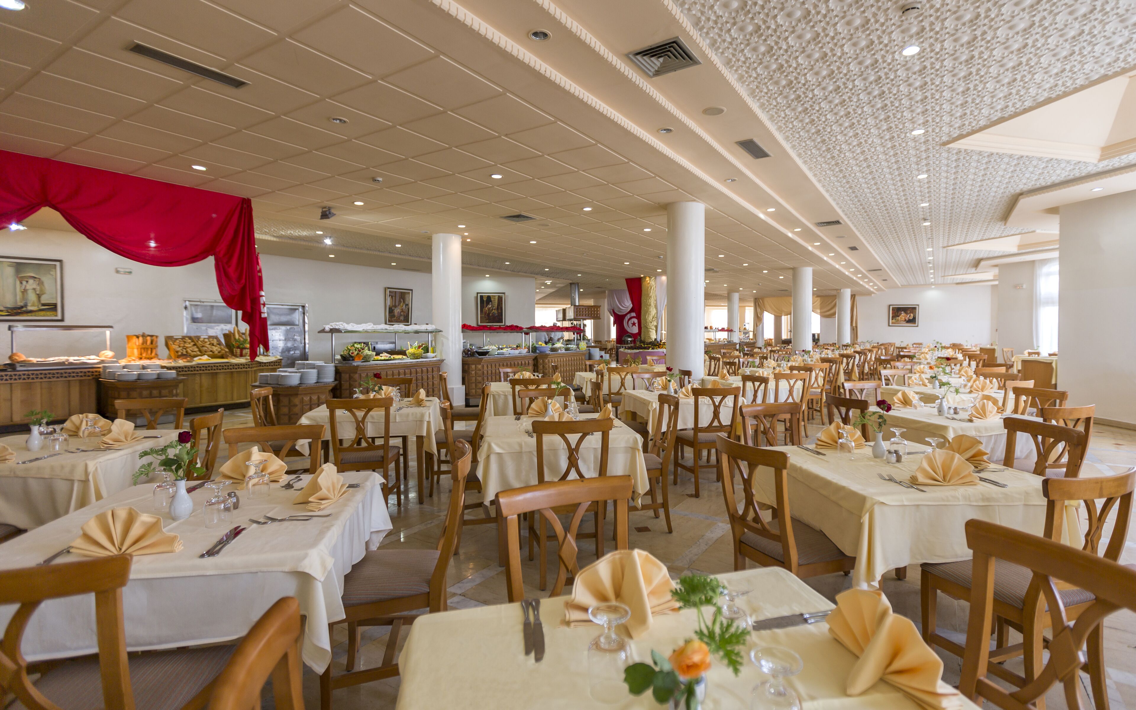 A spacious dining area with multiple tables set for guests, featuring a high ceiling with decorative elements and a buffet area in the background.
