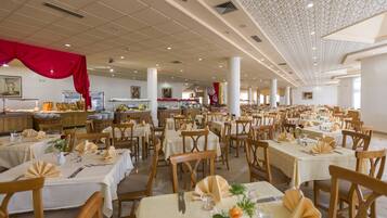 A spacious dining area with multiple tables set for guests, featuring a high ceiling with decorative elements and a buffet area in the background.