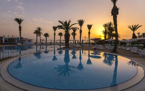 Una piscina de borde circular, rodeada de sillones de descanso y palmeras, con un atardecer de fondo.