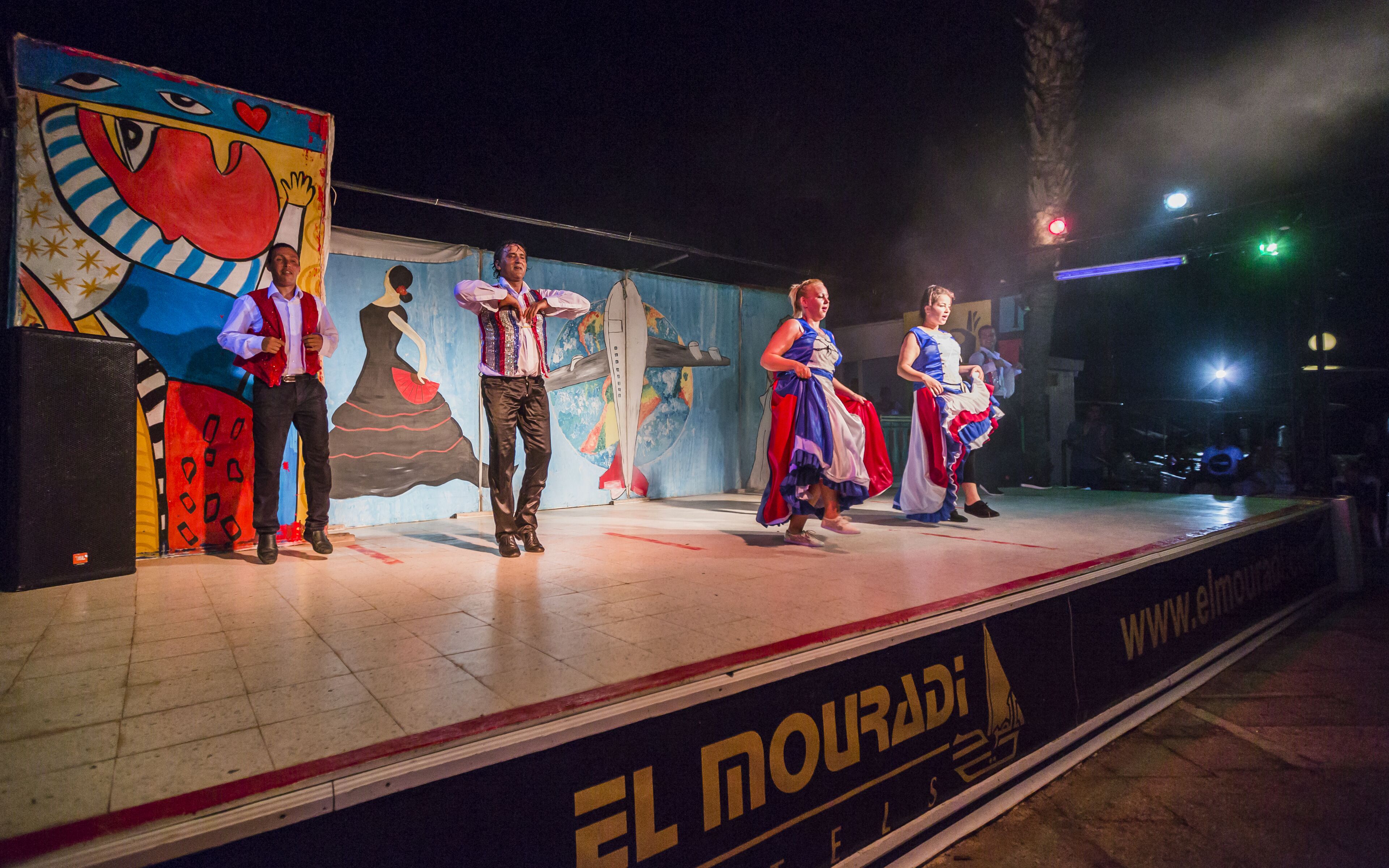 A stage with performers in traditional costumes, a colorful mural, and a banner with "EL MOURADIA" text.