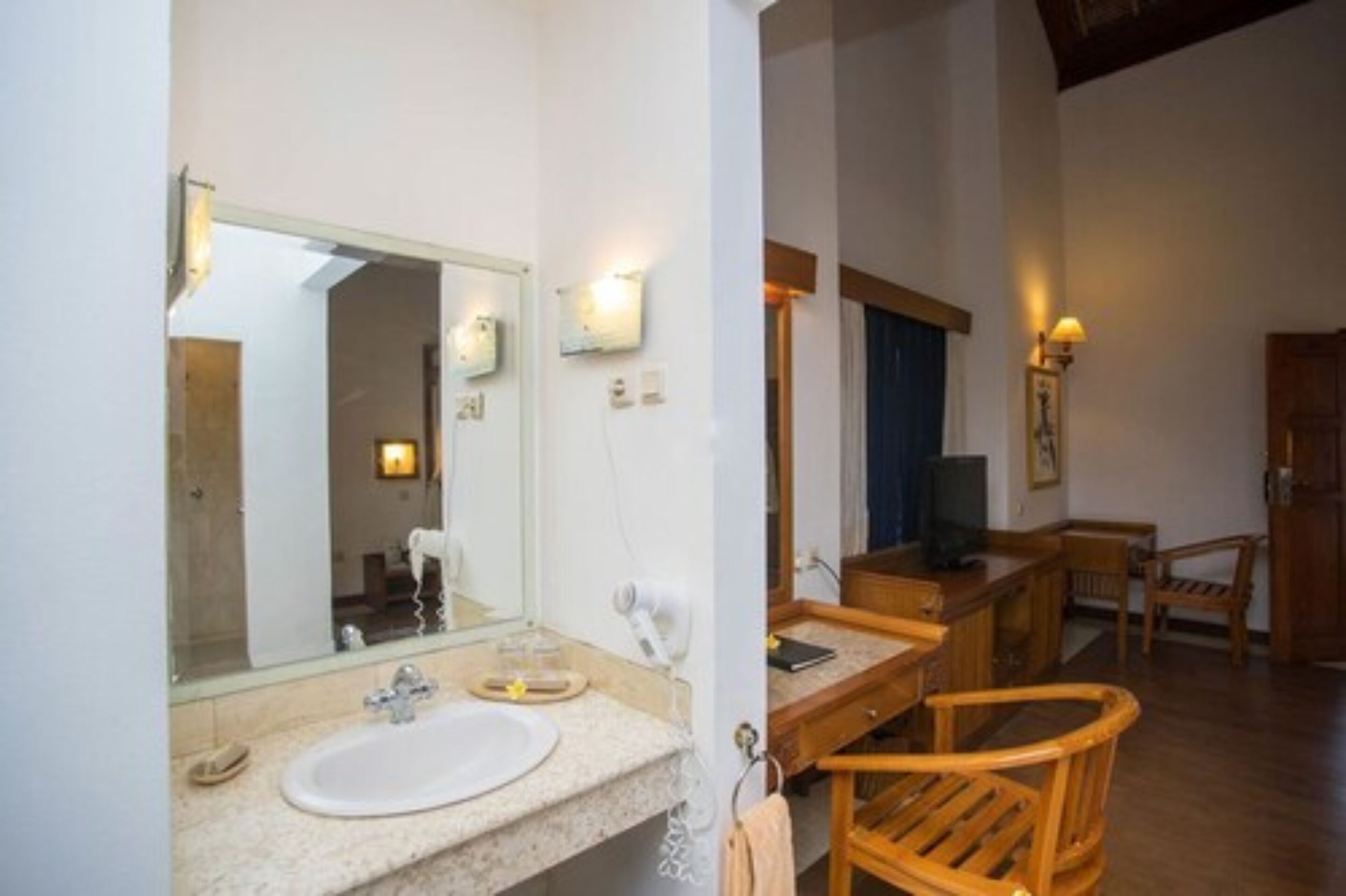 A bathroom with a large mirror, a sink, and a hairdryer. There is a wooden chair and a small table with a television.