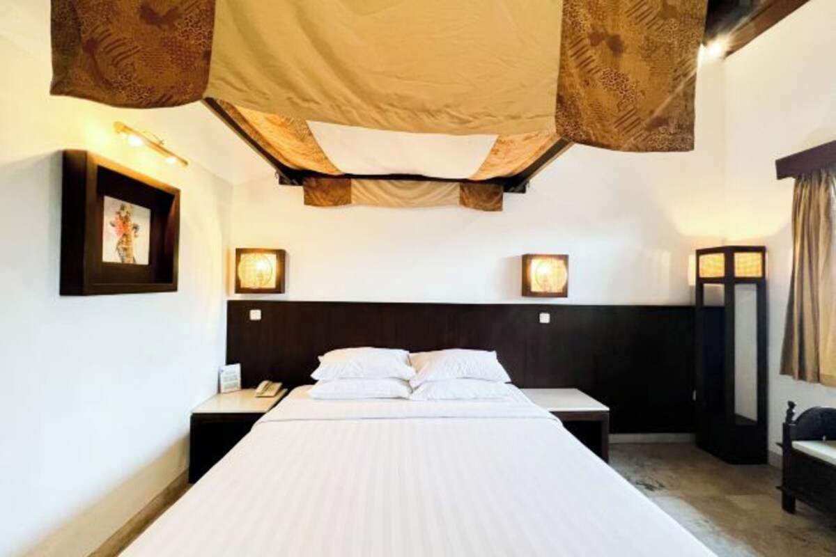 A hotel room with a large bed, two bedside tables, a nightstand, a wall-mounted lamp, and a hanging canopy.