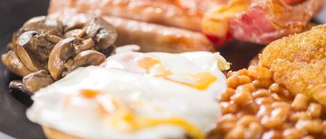 A plate of breakfast food including eggs, sausages, bacon, mushrooms, baked beans, and a fried item.