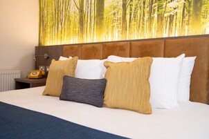 Habitación de hotel con una cama grande, varios almohadones, una mesita de noche con teléfono y una pared con un mural de un bosque.