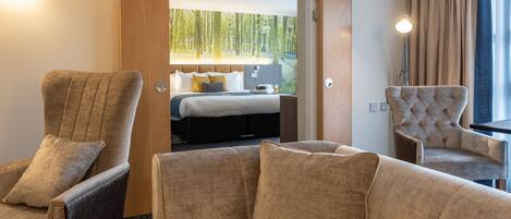 Ein Hotelzimmer mit einem Bett, zwei Sesseln, einem Schreibtisch und einem großen Fenster mit Blick auf einen Wald.