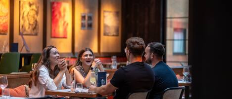 Un grupo de personas cenando en un restaurante con mesas y sillas de madera, obras de arte en las paredes e iluminación cálida.