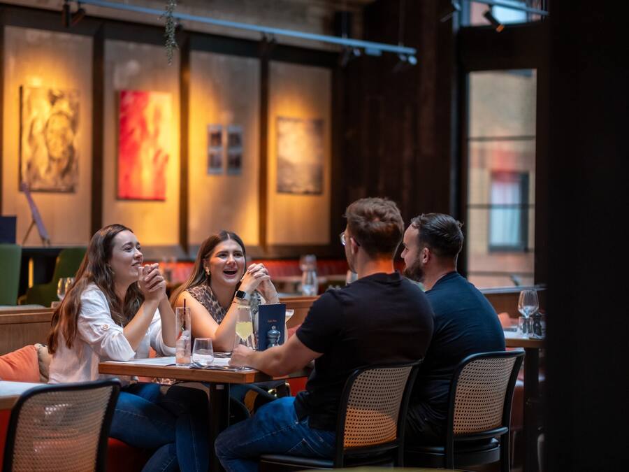Een groep mensen dineert in een restaurant met houten tafels en stoelen, kunst aan de muren en warme sfeerverlichting.